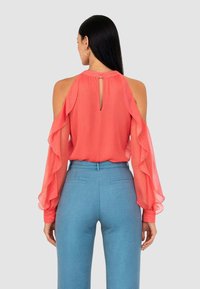 Blusa color corallo con spalle scoperte e maniche arruffate; dettaglio a occhiello sulla schiena. Abbinate a pantaloni azzurri su misura.