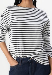 Długi rękaw, w paski, w kolorze białym i granatowym, wykonany z bawełny, z szerokim dekoltem i luźnym krojem. Uzupełniony ciemnoniebieskimi dżinsami.