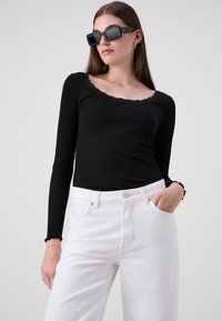 Fekete bordázott, hosszú ujjú felső, fodros dekoltárral, fehér egyenes szárú farmernadrággal párosítva. A modellen fekete négyzet alakú napszemüveg van.