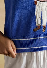 Sinine kootud kampsun valgete triipudega ääres, osaliselt tikitud kujutis beežides kingades, kantud valge tekstuurse seeliku kohal, nähtaval on käsi ja sõrmused.