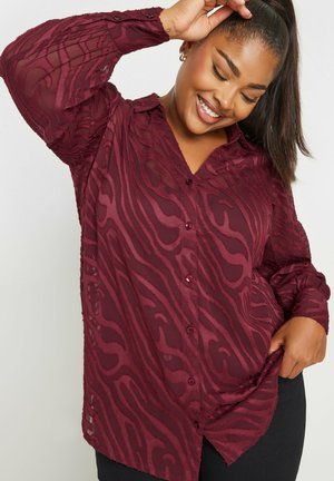 Blusa bordeaux a fantasia in stoffa semitrasparente con chiusura a bottoni, maniche lunghe e motivi ondulati testurizzati. Presenta un colletto a punta.
