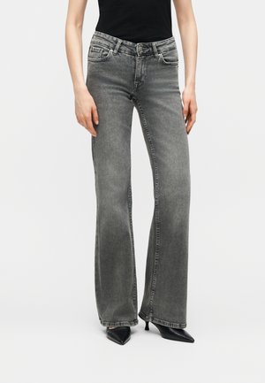 Nő, aki szürke bőszárú farmert és fekete, hegyes orrú, magassarkú cipőt visel, egyszerű fehér háttér előtt áll.