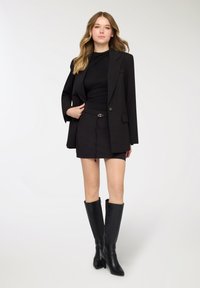 Blazer nero su misura sopra un top aderente nero e una minigonna. L'outfit presenta un tessuto liscio, una vita con cintura e stivali neri alti.