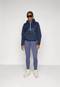 Marine isolert jakke med glidelås, polstret front og elastiske mansjetter, kombinert med lyseblå leggings og beige sneakers. Svarte solbriller brukt.