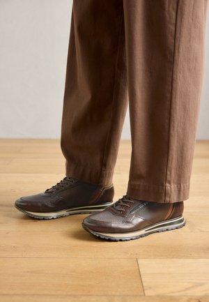 Hnědé kožené tenisky kombinované s volně střiženými hnědými kalhotami na světlé dřevěné podlaze.