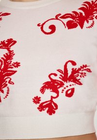 Maglione corto bianco con intricati motivi floreali rossi lavorati a maglia, scollo rotondo e orlo a coste. Tessuto morbido con una vestibilità leggermente oversize.