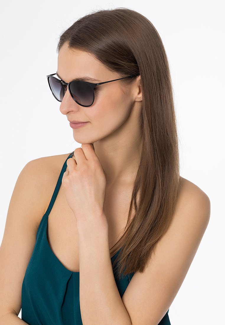 Ochelari de soare cu lentile închise la culoare și un cadru negru elegant, având un design minimalist. Persoana care îi poartă are părul lung și brunet și este văzută din profil.