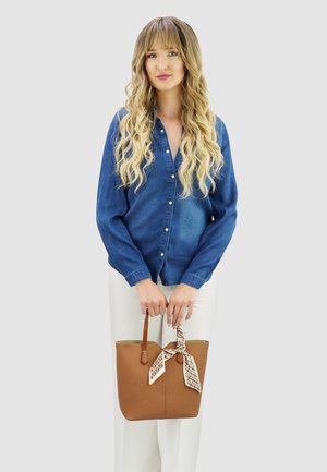 Bruine faux leren tote bag met een gestructureerd oppervlak en een patroon sjaal die aan de hengsel is geknoopt. Het model draagt een blauwe overhemden en witte broek.