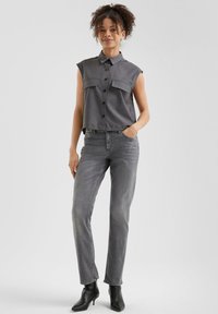 Camisa gris sin mangas con botones en la parte delantera y dos bolsillos en el pecho, combinada con jeans gris de pierna recta y botines negros.