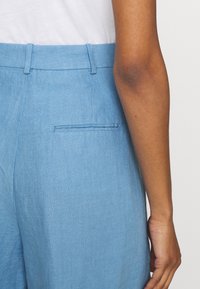 Ljusblå linnbyxor med flat front, bälteslopp och en enda bakficka. Tyget har en slät, texturerad finish.