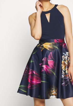 Kvinde iført mørkeblå ærmeløs top med nøglehuludskæring og mørkeblå nederdel med stort, farverigt blomsterprint.