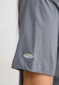 Šedé tričko s krátkým rukávem vyrobené z měkké látky, s reflexní nášivkou loga Nike na levém rukávu. Hladký povrch a pohodlný design.