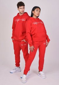 Rode hoodie en joggingsbroek, gemaakt van sweatstof, met een voorzak en witte logo versieringen. Sneakers zijn grijs en wit.