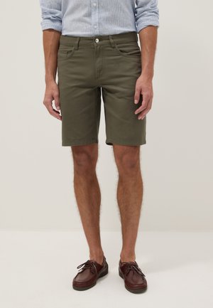 Uomo con pantaloncini verde oliva fino al ginocchio, camicia azzurro chiaro con maniche rimboccate e scarpe stringate in pelle marrone scuro, in piedi davanti a uno sfondo semplice.