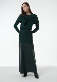 Frau in einem dunkelgrünen Pullover mit einer großen Schleifenapplikation, einem passenden langen Rock mit Gürtel und schwarzen Stiefeletten, stehend vor einem weißen Hintergrund.