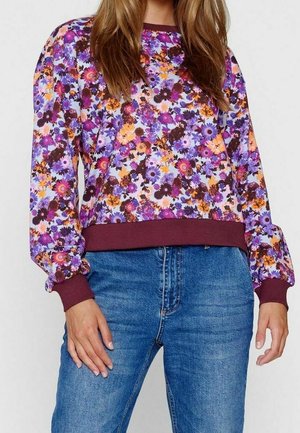 Femme portant un haut à manches longues à motifs floraux en violet, orange et rose avec des poignets et un ourlet marron, assorti à un jean en denim bleu.