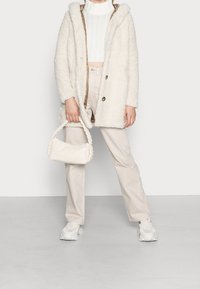 Person som bär en krämfärgad sherpa-kappa, vit kortärmad tröja, beige byxor, vita chunky sneakers och håller en liten off-white flätad handväska.