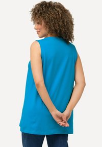 Top senza maniche turchese con vestibilità rilassata, texture liscia, scollatura arrotondata e orlo posteriore arrotondato. Il modello ha i capelli ricci e le mani intrecciate dietro la schiena.