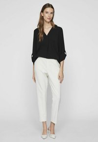 Svart V-ringad topp med rullade ärmar, kombinerad med vita smala byxor. Modellen har på sig ljusa, spetsiga klackar. Enkel och modern design.