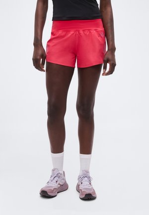 Person trägt leuchtend rote Sportshorts, weiße Kniesocken und pink-graue Laufschuhe und steht vor einem schlichten hellen Hintergrund.