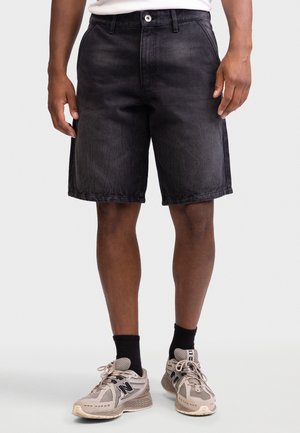 Muž oblečený v čiernych rifľových šortkách, čiernych členkových ponožkách a béžových bežeckých topánkach stojaci na neutrálnom pozadí.