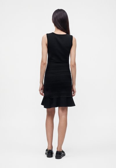 Femme aux longs cheveux foncés portant une robe noire sans manches avec une jupe texturée et un ourlet à volants, debout de dos sur un fond uni.
