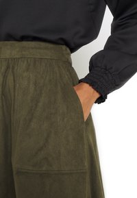 Pantalones de ante verde oliva con cintura alta y bolsillos laterales, combinados con una blusa negra texturizada con puños fruncidos.