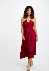 Abito lungo borgogna con corpetto aderente, dettagli arricciati, spalline a halter e orlo fluttuante a volants. Abbinato a sandali dorati.
