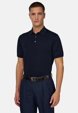 Jeune homme aux cheveux courts portant un polo bleu marine foncé, rentré dans un pantalon plissé bleu marine avec une ceinture en cuir noire.