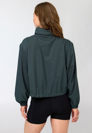 Kobieta o długich brązowych włosach, ubrana w ciemnozieloną kurtkę i czarne spodenki rowerowe, stojąca tyłem na jednolitym tle.