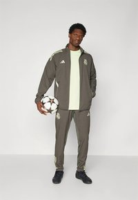 Tracksuit cinzento da adidas com detalhes em verde-claro. O modelo segura uma bola de futebol preta e branca. O design apresenta o logótipo do Real Madrid.