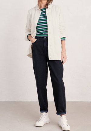 Frau trägt schwarze High-Waist-Jeans, grün-blau gestreiftes Hemd, weiße, geknöpfte Jacke und weiße Sneakers, steht mit einer Hand in der Tasche.