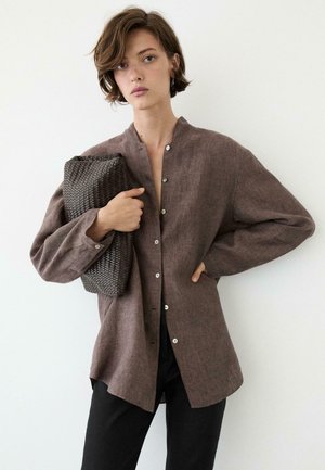 Donna con capelli corti castani che indossa una camicia marrone ampia con bottoni e pantaloni neri, che tiene una grande clutch scura intrecciata contro un muro bianco.