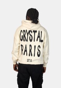 Felpa crema con testo in bold nero che recita "CRYSTAL PARIS" e "DEPT.75" sul retro. Progettata con una vestibilità comoda e cappuccio.