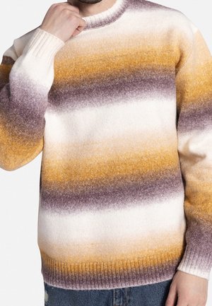 Mężczyzna noszący miękki dziany sweter z poziomymi paskami w kolorach musztardowym, brązowym i białym, w zestawieniu z niebieskimi dżinsami.