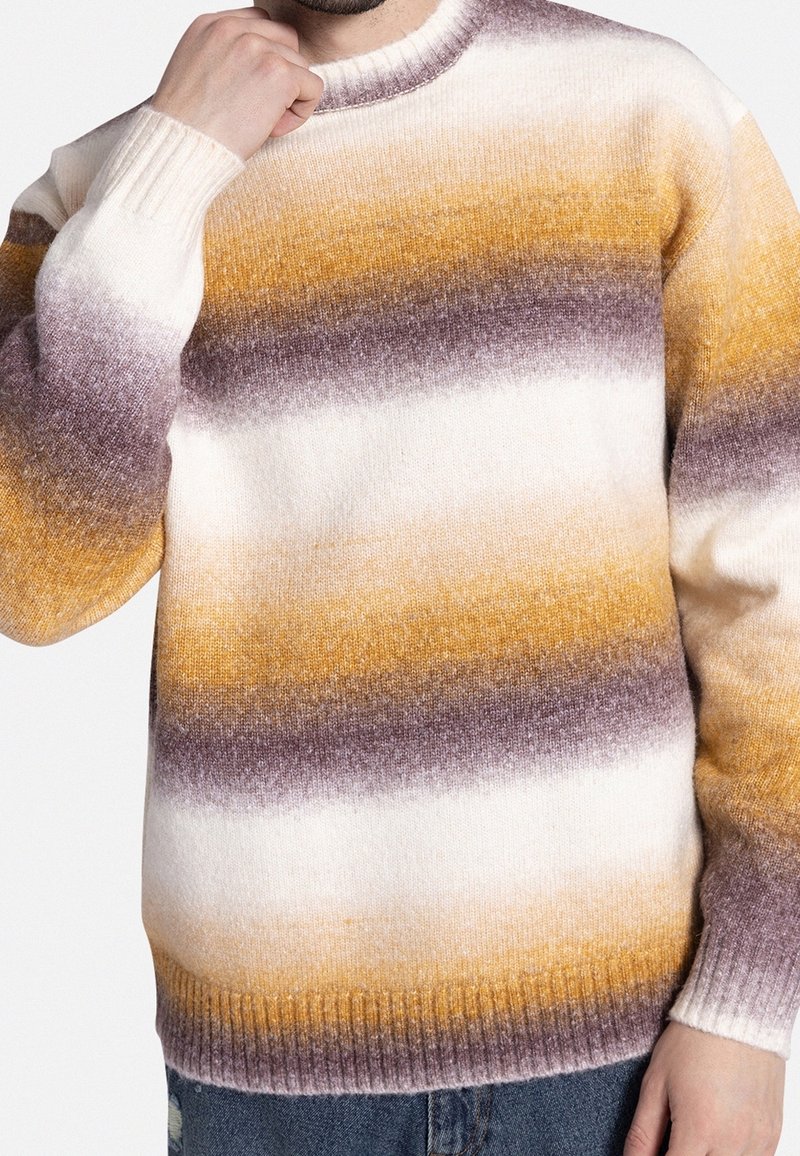 Homme portant un pull en maille douce avec des rayures horizontales jaune moutarde, marron et blanc, associé à un jean bleu.