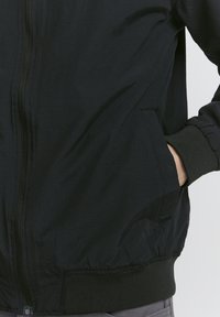 Black bomber jacket made of lightweight material, featuring a front zipper, side pockets, and ribbed cuffs and hem. Smooth texture.