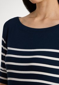 Γυναίκα που φοράει μπλε σκούρο τοπ με οριζόντιες λευκές ρίγες και φαρδύ, στρογγυλεμένο ντεκολτέ που αποκαλύπτει τον λαιμό και το πάνω μέρος του στήθους της.