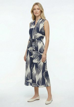 Femme portant une robe midi sans manches bleu marine et beige à motifs avec ceinture à la taille, debout et regardant devant, chaussée de ballerines beiges.