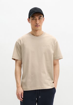 Mann trägt beige kurzärmeliges T-Shirt und schwarze Kappe, steht mit einer Hand in der Tasche vor einem schlichten weißen Hintergrund.