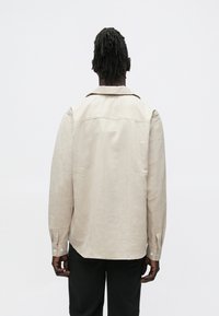 Beżowa koszula z długim rękawem, z kołnierzykiem, mankietami na guziki, teksturowaną tkaniną i subtelnym wzorem. Widok z tyłu ukazuje płaskie szwy i luźny krój.