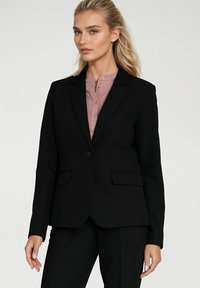 Czarna dopasowana marynarka z dwoma przednimi kieszeniami, z pojedynczym zapięciem na guzik i klapami, noszona na jasno różowej bluzce z kołnierzykiem.