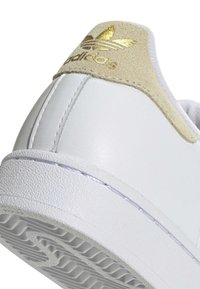 Λευκή φτέρνα αθλητικού παπουτσιού Adidas με μπεζ σουέτ λεπτομέρεια και χρυσό λογότυπο Adidas, δείχνει υφασμένη λευκή καουτσούκ σόλα.