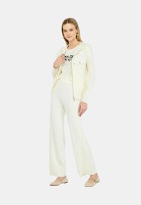 Maglione bianco lavorato a maglia con cappuccio, chiusura con zip e due tasche frontali, abbinato a pantaloni bianchi larghi a coste e ballerine color carne.