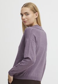 Vijoličasta pletena jopica z dolgimi rokavi in ovratnikom, ki ima mehak material in prilega telo, prikazana na modelu iz stranskega kota.