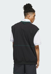 Haut sans manches noir avec des accents verts, présentant une texture lisse, des épaules larges et un ourlet en bande, porté par-dessus une chemise à col blanc.