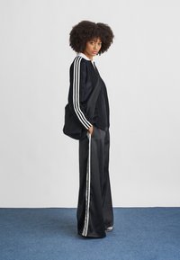 Jachetă din nylon neagră cu dungi albe, combinată cu pantaloni din satin negri care au o dungă albă pe lateral. Include o poșetă neagră de umăr.