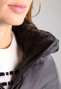 Dark puffer jacket with a brown faux fur collar. Features a zip closure and a white sweater underneath with black stripes and textured pattern.