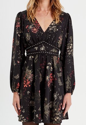 Vrouw draagt een zwarte jurk met lange mouwen, met een rood en beige bloemen- en vlinderprint en een V-hals met een decoratieve rand ter hoogte van de taille.