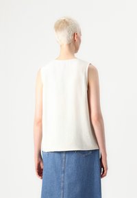 Top sans manches, de couleur beige clair avec une surface texturée ; la vue de dos montre une coupe décontractée et un ourlet droit, associé à une jupe en denim.